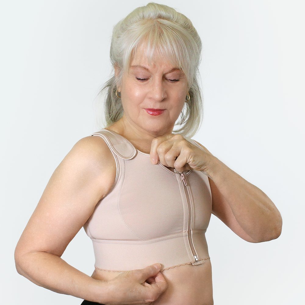Woman zipping up a beige front-zip compression bra with adjustable hook-and-loop shoulder straps